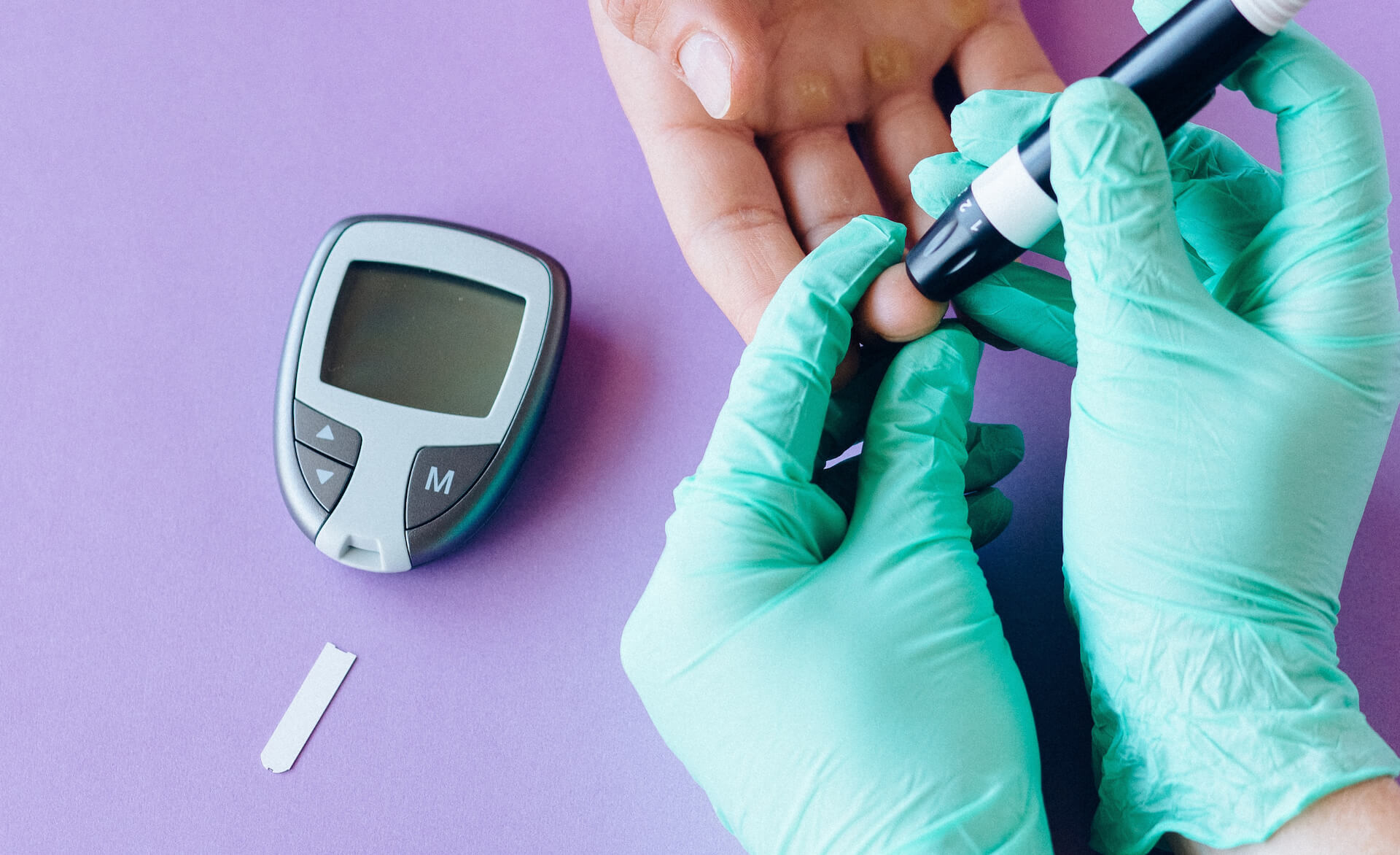 hands wearing green medical gloves holding some finger pricking equipment and pricing the hand of another person