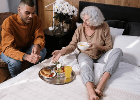 2 people in a bedroom. An older woman is sat on the bed looking at a tray of food. A younger man is sat at the side.