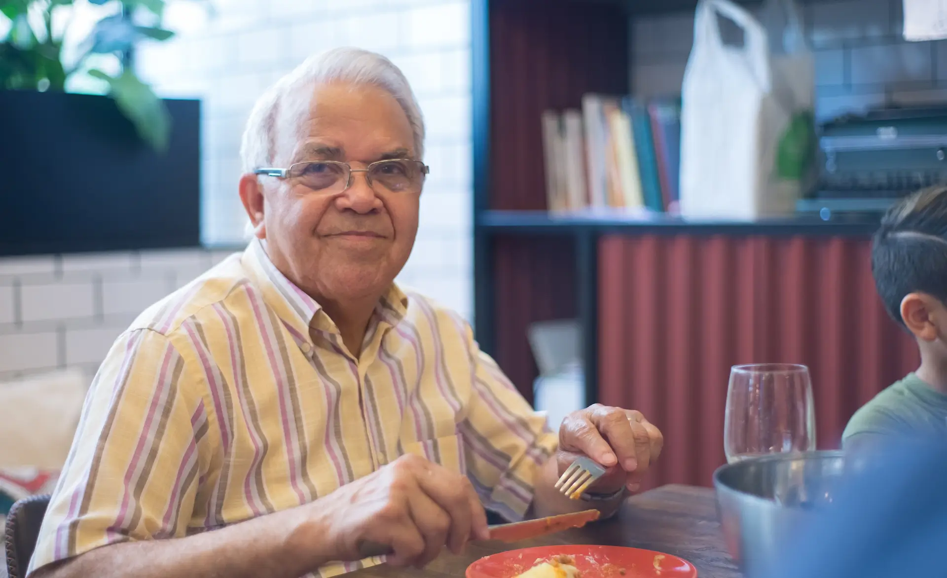 an older gentleman eating a meal while looking into the camera and smiling