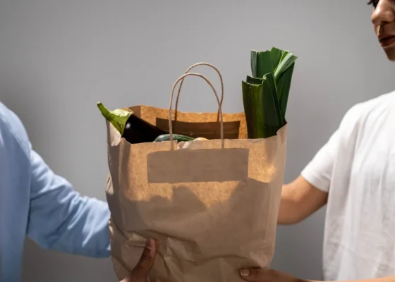 two people holding a brown paper bag with vegetables