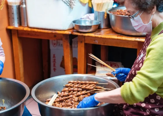 lady stood over a large cooking pot making a type of kebab skewer