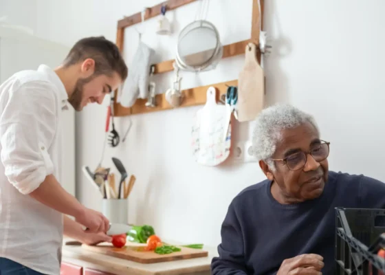 man wearing a navy blue jumper sat at a table but looking over his shoulder. Another man in white is preparing food