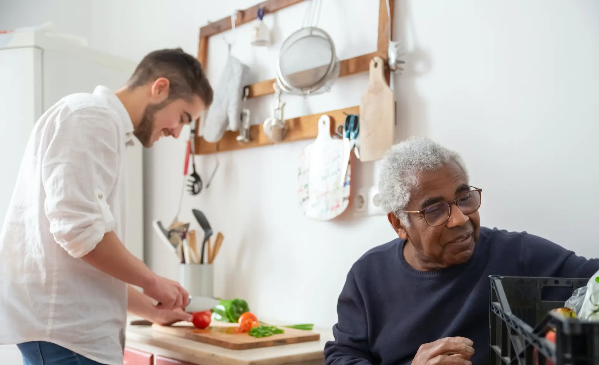 man wearing a navy blue jumper sat at a table but looking over his shoulder. Another man in white is preparing food