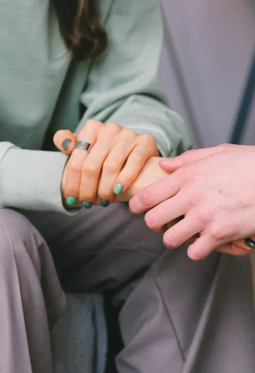 two people sat together. One is wearing hospital scrubs and is holding the hand of the other person