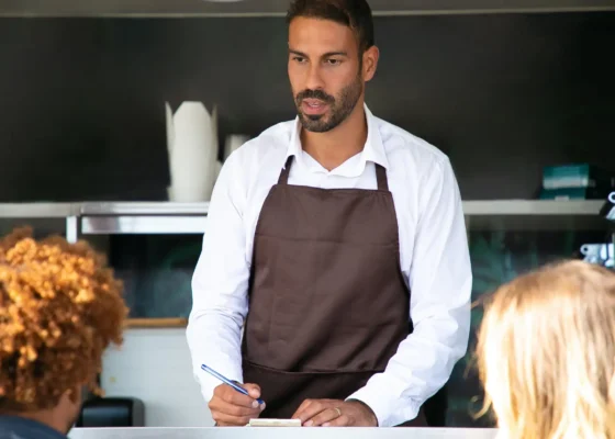 a man wearing chefs whites and a brown apron is holding a pen and writing paper. He is stood at a table.