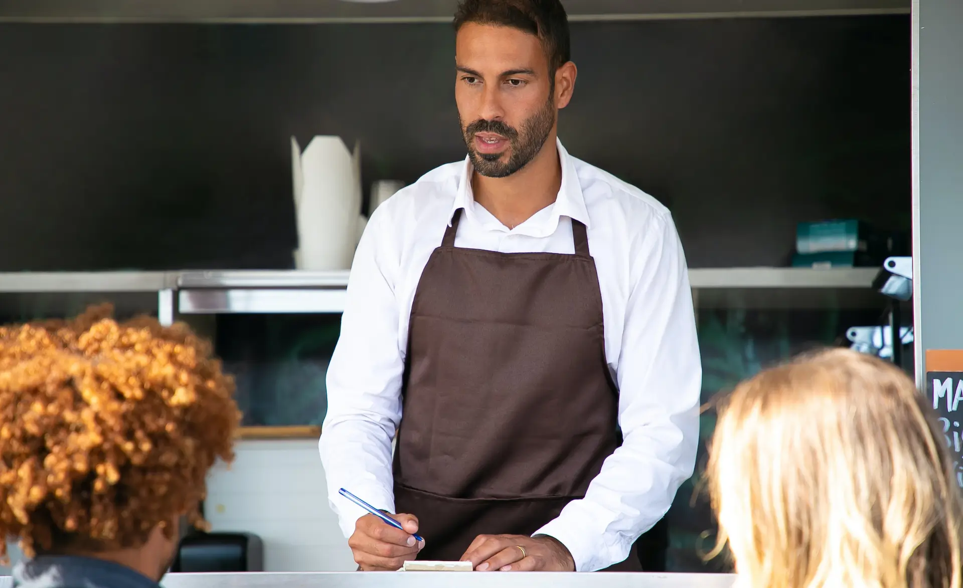 a man wearing chefs whites and a brown apron is holding a pen and writing paper. He is stood at a table.