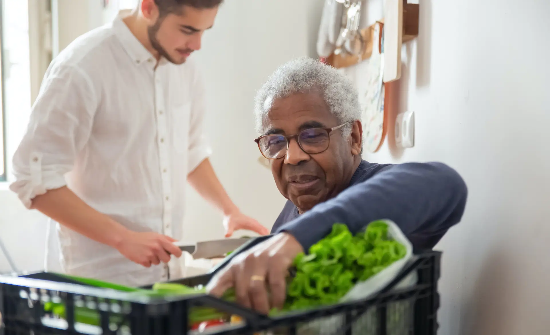 man wearing a navy jumper is holding some leafy green vegetable whilst another man prepares food.